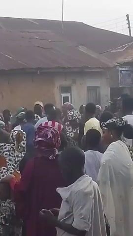 A crowd of people are gathered outside. The scene appears to be in a village or town, with buildings made of mud brick and corrugated metal roofs visible in the background. Many of the people are wearing colorful clothing and headscarves, suggesting a cultural or religious gathering. Some individuals appear to be children. The overall atmosphere seems busy, with many people talking and interacting. The sky is overcast.