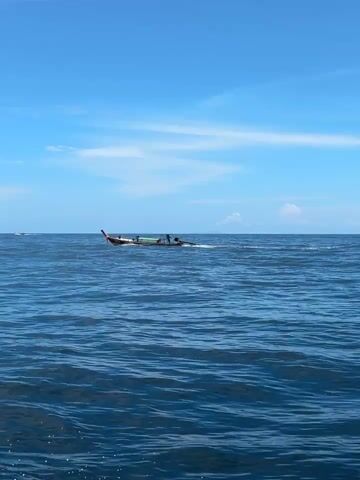 The video shows a vast expanse of deep blue ocean under a clear, bright blue sky with scattered white clouds. The water has small, gentle waves. In the distance, a long, narrow boat with a wooden hull and an engine is visible. Several people are on the boat, appearing to be engaged in some activity. The horizon is clearly defined, separating the sea from the sky, with a hint of land or islands barely discernible in the far distance. The camera appears to be mounted on another moving vessel, as evidenced by the slight movement and the perspective of the water.