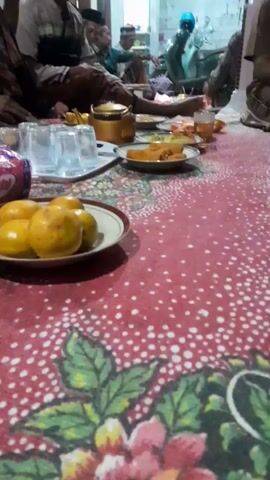 A group of men are seated on the floor, around a low table laden with food and drinks. The floor is covered with a red and white polka dot patterned cloth, with floral motifs along the edges. In the foreground, a plate of oranges is visible. Further back, there are plates of fried food, glasses, and a golden teapot. The men are dressed in traditional clothing, with some wearing caps. One man in the background appears to be holding a phone to his ear. The environment appears to be indoors, possibly a home, with a wall and a doorway visible in the background.