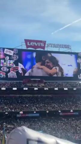 The video shows a view of Levi's Stadium, packed with a large crowd of people. A massive screen displays a series of images and videos, transitioning from a couple embracing amid confetti, to a football game scene, and finally, a close-up of a football player. The sky is blue with a contrail visible. The stadium's name, "Levi's Stadium," is prominently displayed on the structure above the screen. Banners and logos are visible around the screen and within the stadium.