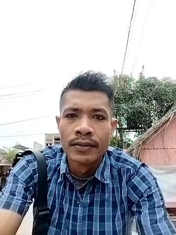 A man in a blue and black plaid shirt and a shoulder bag is sitting on a motorbike, filming himself. The motorbike is stationary. He is facing the camera, with a wooden shack and trees behind him. The shack is painted pink. The day is overcast.
