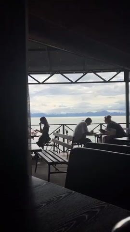 This video shows a view from inside a dimly lit, covered outdoor seating area, likely a restaurant or cafe, looking out towards a body of water and mountains. In the foreground, dark upholstered seats and wooden tables are visible. A metal railing separates the seating area from the water. Beyond the railing, the water stretches out to meet a distant shoreline with snow-capped mountains under a cloudy sky. Three people are seated at tables overlooking the water. On the left, a person is seated alone at a small table, appearing to be looking down at something. In the middle, a man and a woman are seated at a table. The man is looking down at his hands or the table. The woman is looking towards the man and gesturing with her hands. They appear to be engaged in a conversation. Wooden benches are also present in the seating area.