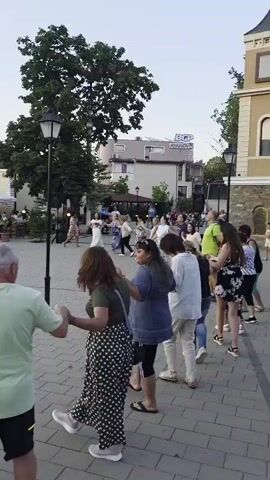 The video shows an outdoor gathering of people dancing in a square. The camera focuses on a group of people in the foreground dancing in a circle, holding hands. They are all facing inwards and appear to be moving to music. In the background, more people are visible, some dancing, others sitting at tables outside a building that has umbrellas providing shade. There are trees and lampposts in the square, and the sky is visible overhead. The lighting suggests it is either early morning or late afternoon. The overall atmosphere seems festive and communal.