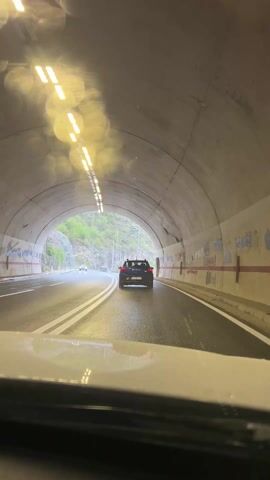 The video captures a first-person perspective from inside a car driving through a tunnel and then out into a rainy, mountainous landscape in Dobrsko selo, Montenegro.

The initial shot is from within a concrete tunnel with a rounded arch, illuminated by bright linear lights on the ceiling. The road surface is visibly wet. A black SUV is directly ahead, followed by a lighter-colored car further down the tunnel. The right wall of the tunnel is covered in various red and black graffiti, including symbols and partially visible text such as "KM/H".

As the car exits the tunnel, the scene transitions to an overcast and rainy day, with a misty atmosphere engulfing the surrounding rocky hills and sparse green vegetation. The black SUV ahead has been replaced by a blue SUV, whose Montenegrin license plate begins with "MN". The wet asphalt road is flanked by streetlights. The video concludes with the car continuing its journey along the winding road amidst the misty, mountainous terrain.