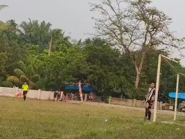 A soccer game is in progress on a grassy field. In the foreground, two players in striped uniforms are visible. One player is dribbling the ball, while the other player is standing near the goal. In the background, spectators are watching the game from under a blue tent, and lush green trees and foliage surround the field. The sky is overcast.