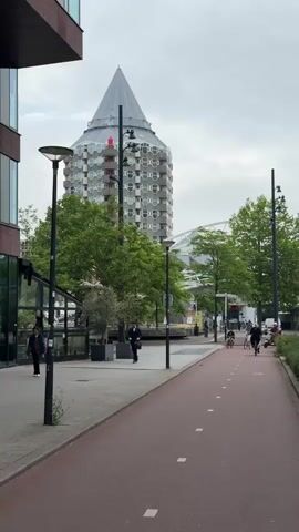The video shows a paved path with a red surface marked with white dashes, indicating a bike lane. Several people are riding bicycles and a scooter on the bike lane. In the background, there is a tall, distinctive building with a pointed roof and many windows. Green trees are visible throughout the scene, and the sky is overcast. To the left of the bike lane, there is a sidewalk with lampposts and some people walking. The overall environment appears to be an urban setting with a focus on pedestrian and cyclist infrastructure.