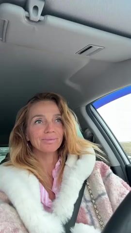 The video depicts a woman with shoulder-length strawberry blonde hair sitting in the driver's seat of a car. She is wearing a pink and white striped shirt, a gold necklace, and a pink and white plaid coat with a white fur collar. The car's interior is visible, including the dashboard, sun visor, and seatbelt. The car is moving. The background shows a glimpse of a bright, possibly overcast sky. Music is playing.
