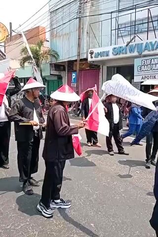 A procession is moving down a paved street under a clear sky. Several individuals, mostly men, are walking in single file. They are all wearing conical hats with red and white bands. They are also wearing black long-sleeved shirts and black pants. Some of them are carrying flags, predominantly red and white, some are on poles, while others are held by hand. The flags appear to be Indonesian flags. Some individuals are holding short wooden sticks. In the background, there are buildings lining the street, one of which is a pharmacy with a sign reading "APOTHIK SRIWIJAYA". There are also spectators on the side of the street. A vehicle, possibly a float, with the words "SENTRA PENGEMBANGAN IKAN DEMAK" on it is visible further down the procession. The overall atmosphere seems festive.