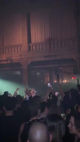 The video depicts a crowded indoor venue, likely a concert or nightclub, filled with people dancing and raising their hands in the air. The space has an elevated balcony. Strobe lights flash in various colors, illuminating the scene. Some attendees are recording with their phones.  A close-up shot shows a man with curly hair and a brown cap looking at the camera. The camera then pans back to the crowd.