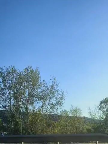 The video shows a view from a vehicle moving along a road. The sky is clear and blue. On the right side of the frame, there are several trees, some with green leaves and others bare. In the distance, rolling hills are visible with some buildings clustered on them. A metal railing or barrier runs along the bottom of the frame, suggesting the perspective is from a highway or elevated road. The video captures the movement of the vehicle by showing the landscape passing by. At one point, a light-colored van and a car are seen on the road ahead, indicating traffic. The lighting suggests it is daytime, likely morning or afternoon, with the sun casting a bright light.