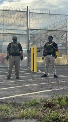 The video shows two security officers standing in front of a fence topped with barbed wire. The officer on the left is wearing a helmet, a tactical vest labeled "Police", and a patterned face covering. The officer on the right is wearing a tactical vest labeled "ERO" and a patterned face covering. Both officers wear gloves and khaki-colored pants with black shoes. In the background is a fence with a sign that reads "Gate 5, Caution Speed Bump." The ground is asphalt, with some vegetation in the foreground. In the background, the sky is visible with some cloud cover.