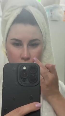 In a medium shot, a woman with fair skin and dark hair wrapped in a white towel applies a clear product to her face while holding a black smartphone in her left hand. The phone's case is adorned with small, embossed hearts and the camera lenses are clearly visible. Her nails are painted a light purple. She is looking directly at her reflection in the phone's screen with a focused expression. The background is slightly blurred, revealing a bathroom setting with a white cabinet that has various personal care items on top.