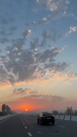 The video shows a view from inside a car driving on a highway at sunrise. The sun is low on the horizon, casting an orange glow across the sky. There are scattered clouds in the sky, some illuminated by the sun's rays. The road is paved and has white dashed lines marking the lanes. On the left side of the road, there are trees and a guardrail. On the right side of the road, there is also a guardrail. A black car is driving in front of the camera car. The overall scene depicts a peaceful and scenic sunrise drive.