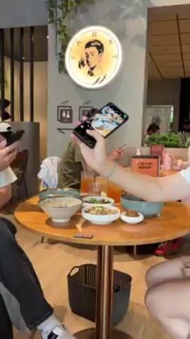 A hand is holding a smartphone, taking a photo of several bowls of food and drinks on a round wooden table in a restaurant. Another person is in the background, also looking at a phone. Restaurant decor includes signs for restrooms, a hanging lamp with a picture, and a sign in Chinese.