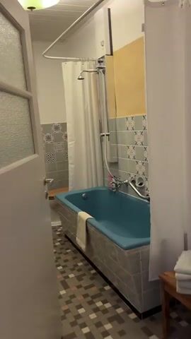 The video opens with a brief shot of a bathroom. The camera then pans to a bedroom where a man is lying on a bed looking at his phone. He is wearing a dark t-shirt and light-colored pants. He is on his stomach, propped up on his elbows. To the right of the bed, a woman is sitting in a chair, also looking at her phone. She is wearing a dark jacket and appears to be on her side. The room contains a bookshelf, a lamp, a decorative palm tree, a window with curtains, and a small table with some items on it. A bottle of water is on the floor near the bed. The lighting in the room suggests it is either early morning or late evening. The overall impression is of a relaxed domestic scene.