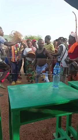 The video shows a man dancing in front of a crowd. He is wearing a black crop top and jeans with patches. In the background, a crowd of people, including some dressed in traditional clothing, watches him. A man in the crowd holds what appears to be a bundle of animal hair. The dancing man waves his arms, kicks his leg, and gyrates his body. A green table with a bottle of water is in the foreground. The setting appears to be outdoors, in a field.