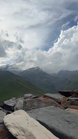 The video is a vertical shot of a mountain landscape under a cloudy sky. In the foreground, there are large, rough rocks with hints of orange and white coloration. Beyond the rocks, rolling green hills lead to jagged, snow-capped mountains in the distance. The sky is filled with dramatic clouds, with patches of sunlight breaking through. A winding road is visible on one of the green hills. The overall impression is a majestic and somewhat wild natural environment.