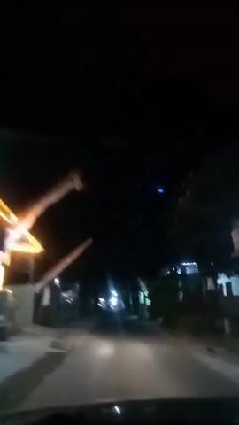 The video shows a first-person perspective from inside a vehicle driving at night on a road lined with buildings and vegetation. The headlights of the vehicle illuminate the road ahead. There are lights visible from the buildings, some of which appear to be shops or houses. The sky is dark. There are no visible people or animals. The road surface appears to be asphalt.