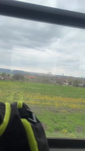 A view of a landscape from inside a moving vehicle, likely a train or bus, is captured. The window frame is visible at the top and sides, with a partial view of a black backpack with neon green accents in the lower left corner. Outside, a field of green grass with many yellow wildflowers extends into the distance. The field leads up to a row of small houses or buildings, surrounded by trees with bare branches, suggesting it may be early spring. In the background, a faint outline of hills can be seen under a cloudy, overcast sky. The scene evokes a sense of travel through a rural or countryside area.