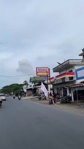 This vertical video captures a street scene in Uteungeulinggang, Indonesia, likely in the late afternoon or early evening given the overcast sky and the muted lighting. The weather appears cloudy, with a significant cloud formation dominating the upper part of the frame.

The main subjects are the various vehicles and people navigating a moderately busy road. Numerous motorcycles are visible, carrying one or more passengers, some wearing helmets and traditional head coverings. A few cars and a small truck are also present, contributing to the traffic flow. The activity is the everyday movement of people and goods along a commercial street.

The setting is characterized by a row of shops and buildings lining the right side of the road. Prominent among these is an Alfamart store, indicated by its distinctive red and white signage. Other businesses include what appears to be a barber shop labeled "JAZ BARBER AND SHOP." The buildings are a mix of styles, some appearing older with corrugated metal roofs, while others are more modern. There are also flags and banners displayed, one large white flag with red lettering, suggesting a local event or promotion.

Notable details include the presence of street vendors or small stalls set up in front of some of the shops, and numerous parked motorcycles. The overall emotion conveyed is one of daily life and commerce in a bustling Indonesian town, with no indication of urgency or distress. The road surface is asphalt, and electrical wires crisscross the sky above the street, typical of urban or semi-urban environments in the region.