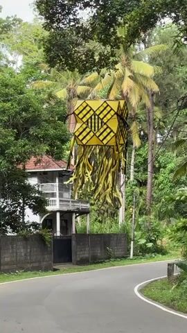 The video presents a static shot of a street scene in Thihagoda, Sri Lanka, on an overcast day. A large, square-shaped yellow lantern, adorned with black geometric patterns and long, tattered yellow streamers, hangs suspended from overhead wires in the center of the frame. Lush green foliage, including tall palm trees, forms the backdrop.

An asphalt road curves gently to the right in the foreground. A red and black Piaggio rpe auto-rickshaw (tuk-tuk) drives away from the camera. The rear of the tuk-tuk displays "STOP" on a red rectangle, "Don't worry Be Happy," and four playing card aces (hearts, diamonds, clubs, spades), along with the identifier "551H." A white Toyota Prius follows the tuk-tuk, also driving away from the camera.

To the left of the road, behind a gray concrete wall, a two-story white house with a balcony on its upper level is partially visible amongst the trees. The scene captures a tranquil, verdant setting characteristic of a tropical region.