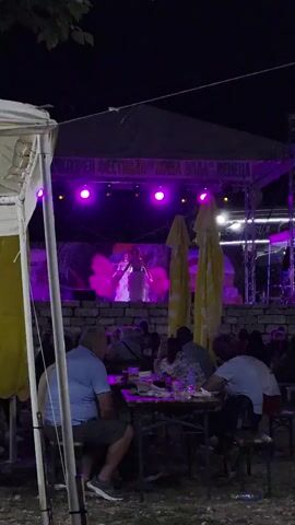 A night-time outdoor performance is taking place on a raised stage. A female performer in a white costume with pink feather fans is dancing in the center of the stage. The stage is lit with purple and orange lights. Behind the performer, a projection screen displays an image of what appears to be tropical foliage. A banner across the top of the stage reads "KULTOREN FESTIVAL "ZIVA VODA" VENECIA". In the foreground, an audience is seated at tables, watching the performance. Several large yellow umbrellas are visible behind the audience. The overall atmosphere appears to be festive.
