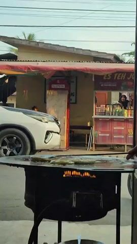 A white SUV drives by a food stall. A person is visible behind the counter of the stall, preparing food. There is a large, round griddle with flames underneath it in the foreground. The stall has red trim and signage, and a red telephone booth is visible behind it. The sky is overcast.