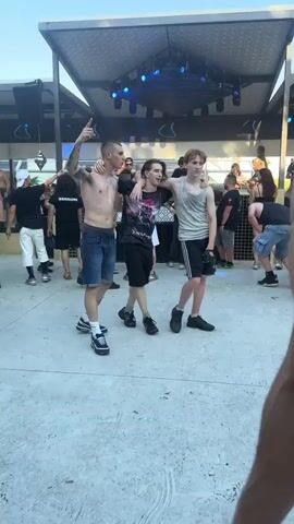 A shaky, first-person perspective video shows a group of young people at an outdoor event. The first frame focuses on three young men posing for a picture, with two of them embracing. The man on the left, who is pointing upwards, has tattoos on his arms and is wearing a denim vest and shorts. The other two men are also casually dressed. In the background, a stage with blue lights is visible, suggesting a concert or performance. Other people are milling around the area. The video then transitions to a close-up of a young man's face as he speaks directly to the camera, with a blurry crowd and smoke in the background. He appears agitated and is speaking in Italian. The final frame is an extreme close-up of a blurred, light-skinned surface, possibly a face or hand, with a dark, out-of-focus object in the corner. The audio consists of spoken Italian phrases, including "Porco Dio" repeated multiple times.