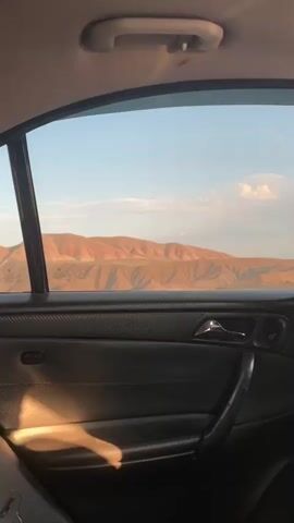 The video is filmed from inside a car, looking out the rear passenger window. The window frame and part of the car's interior door panel are visible in the foreground. The door panel is dark colored and appears to be leather or a similar material, with a chrome door handle and window control. Sunlight is casting a shadow across the door panel. Outside the window, a mountainous landscape is visible under a clear blue sky with a few wispy clouds. The mountains are arid and appear to be a reddish-brown color, with shadows highlighting their contours. The sun is low in the sky, suggesting it is either early morning or late afternoon. There are no people or animals visible in the frame. The car appears to be in motion, as the landscape outside is a static view.