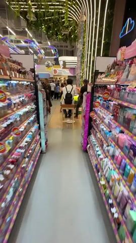 The video shows the interior of a brightly lit store, possibly a convenience store or a gift shop, with shelves stocked with various colorful items. In the foreground, there are two aisles of products, with the camera positioned in the middle, looking down the length of the store. The ceiling is adorned with artificial greenery and curved LED lighting. In the middle ground, several people are browsing or interacting with displays. Most notably, there is a tall, cylindrical display filled with colorful candy or toys, resembling rocket ships, positioned near the center of the store. A few people, including children, are gathered around this display. One person is sitting on a stool, facing away from the camera, wearing a backpack. In the background, more people are visible, and the store appears to have a whimsical or playful aesthetic, with decorative elements like colorful rings hanging from the ceiling.