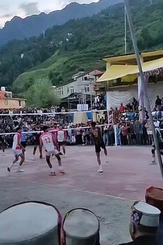 This is a video of a volleyball game being played outdoors in a village. There is a volleyball net set up in the middle of a court. There are players on both sides of the net, wearing jerseys and shorts. The game is being watched by a large crowd of spectators who are standing behind a barrier. The court is surrounded by buildings and trees, with mountains in the background. The sky is cloudy. There are two drums in the foreground, one with a red and white pattern, and the other with a darker pattern.