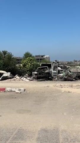The video begins with a wide shot of a junkyard filled with damaged and burned vehicles under a clear blue sky. The foreground is a dusty, uneven dirt road. In the middle ground, several vehicles, including cars and vans, are piled up, some appearing completely destroyed. Green foliage and palm trees are visible behind the wreckage, along with some buildings in the background.

As the video progresses, a person wearing a dark cap and dark clothing walks into the frame from the left, partially obscuring the view. The camera then pans slightly to the right, continuing to show the junkyard. Another person, dressed in a dark shirt and blue jeans, walks across the foreground from left to right, their back to the camera. The junkyard remains the central focus, with more damaged vehicles and debris visible. A person is seen walking in the distance within the junkyard area. The video concludes with a similar view of the junkyard, emphasizing the extensive damage and the desolate environment.