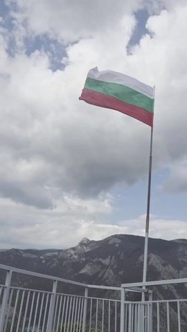 The video shows a Bulgarian flag waving on a pole against a backdrop of mountains and a cloudy sky. The flag is the traditional white, green, and red horizontal tricolor of Bulgaria. It is attached to a metal pole with some wear. The pole is situated behind a white metal railing, suggesting a viewing platform or overlook.  In the background, mountains stretch across the horizon, partially covered in vegetation. The sky is filled with white and gray clouds, giving a slightly overcast appearance.