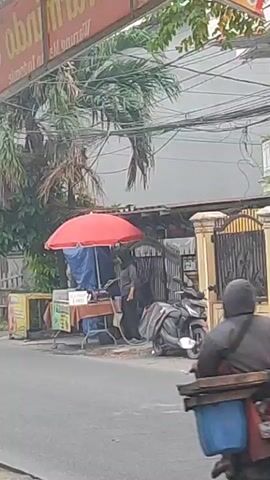 A person wearing a gray jacket and a helmet is riding a scooter down a paved road. The scooter has a large wooden rack attached to the back, filled with blue containers. A red umbrella is propped up above a food stall on the right side of the road, where another person in dark clothing appears to be working. A motorcycle is parked behind the food stall. The road is lined with buildings and trees, and there are many electrical wires overhead. The scene appears to be a street in a tropical or subtropical urban area.