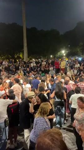 A crowd of people of mixed ages and ethnicities are gathered outside at night, either dancing or watching a performance. Some are standing and moving in what appears to be a dance, while others are seated on tiered steps behind them, watching the activity. Most of the people are dressed casually. There is a tall, thin monument or tree trunk visible in the background, with trees and streetlights further behind. The overall atmosphere is lively and festive.