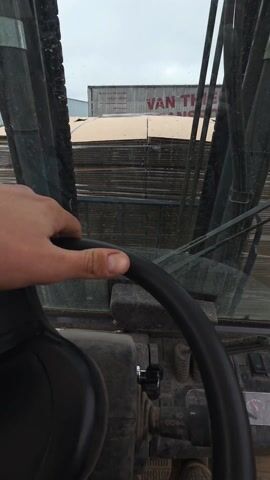 A first-person perspective from inside the cab of a forklift shows a hand on the steering wheel. Through the dirty windshield, large stacks of cardboard are visible. To the left, another forklift is parked. A truck with the word "VAN" on its side is partially visible in the background. The scene appears to be an outdoor storage or industrial area.