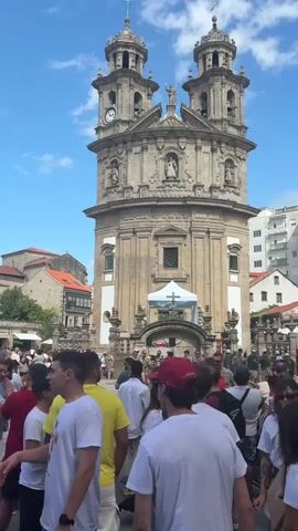 A large stone church with two prominent towers, a clock on the left tower, and a statue on the peak of the facade is the central focus of the video. The church has an ornate, baroque architectural style. In front of the church, a crowd of people are gathered. Many individuals are wearing casual clothing, including t-shirts and shorts. Some people are holding cell phones and appear to be recording or taking pictures. The atmosphere suggests a public event or gathering. The environment appears to be an urban setting with buildings visible in the background, including apartment buildings. The sky is blue with some clouds, indicating daylight. The ground is paved. There are some awnings or temporary structures set up near the church entrance. The overall impression is a lively, outdoor event in front of a historic landmark.