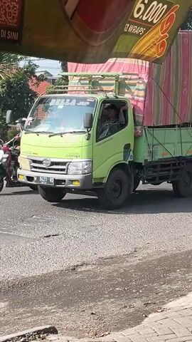 A truck is parked on the side of a road. A person on a motorcycle is driving past the truck. There are other vehicles and trees in the background. The road appears to be paved with asphalt. A sidewalk is visible in the foreground.