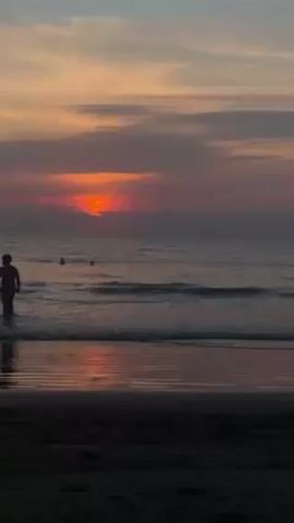 The video shows a beach scene at sunset. The sun is a bright orange-red, partially obscured by clouds. The sky has bands of orange, pink, and gray. The ocean is dark, with gentle waves coming to shore. The beach sand is dark, and reflects the colors of the sunset. There is a person walking in the shallow water on the left side of the frame, and two other people further out in the ocean.