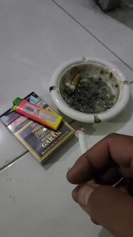 A human hand holds a lit cigarette with a glowing red tip near a white ashtray filled with cigarette butts and ash. A pack of Gudang Garam cigarettes and a pink and yellow lighter lie on a tiled floor.