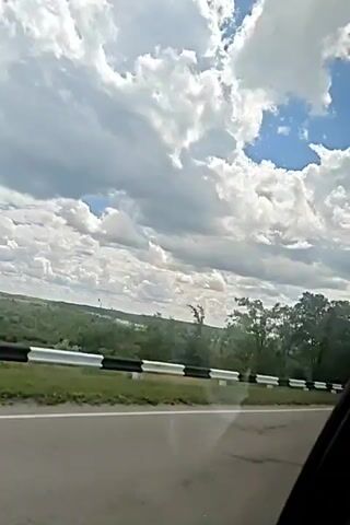 The video shows a car's perspective as it travels along a paved road. A guardrail with black and white stripes runs alongside the road, separating it from a grassy area and then a wooded landscape. In the background, rolling hills are visible under a bright blue sky filled with large, fluffy white clouds. Several trees are scattered throughout the landscape, some appearing bare-branched. The overall impression is that of a scenic drive through a rural or natural environment on a clear, possibly summer day.