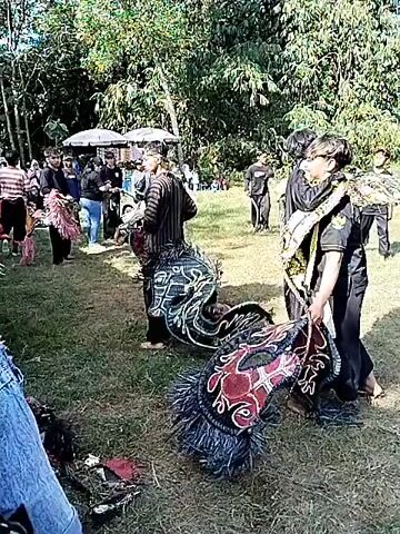 A group of people are gathered in an outdoor grassy area with trees in the background. Several individuals are wearing traditional attire, some with ornate headdresses and patterned garments. One person is on the ground, appearing to be in a prone position. Another individual in the foreground is wearing a black outfit and holding what looks like a decorative fringe. In the middle ground, two people are embracing, one wearing a black outfit and the other with a patterned garment and a headband. In the background, there are tents and more people observing. The overall impression is of a cultural performance or gathering.