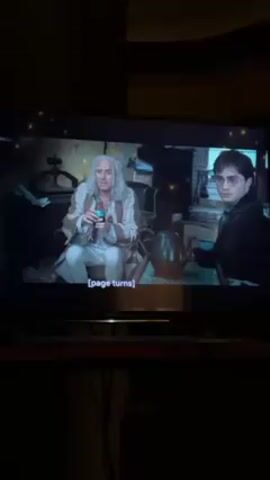 The video shows a television screen displaying a scene from a movie, likely a Harry Potter film. In the initial shot, a man with long, gray hair is seated and holding a potion, while Harry Potter stands nearby. The scene then shifts to another room where Ron Weasley sits near a silver teapot, and Hermione Granger is reading a book. Subtitles on the screen read "There were once three brothers." The environment appears to be a dimly lit, cluttered interior with posters on the walls.