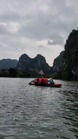 The video shows a body of water, possibly a lake or river, with several small boats occupied by people. The people are wearing orange life vests, and some are rowing the boats. In the background, there are lush green hills or mountains under a cloudy sky. The overall scene suggests a tourist activity or recreational outing on a scenic waterway.