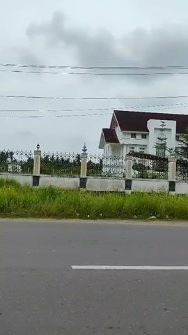 The video shows a white building with a domed roof, a tiled roof, and columns. The building is surrounded by a fence and grass. The sky is cloudy. The video is taken from a car moving down a road.