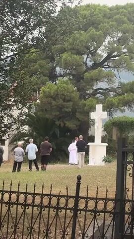 A group of five people are standing in a grassy area in front of a white cross with a sculpture of Jesus on it. Two individuals, one in a white robe and another in dark clothing, stand close to the cross, appearing to be performing a ritual or ceremony. The other three individuals stand further back, observing the event. The environment includes lush green trees and foliage, with a glimpse of a building in the background. A black metal fence is visible in the foreground.