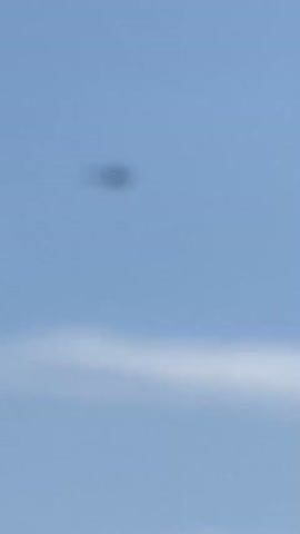 This video captures a clear blue sky with wispy clouds. The primary subject is a helicopter, visible as a small, dark silhouette against the sky. The helicopter appears to be in flight, with its rotors spinning. In the first frame, a blurry, dark circular object is visible, but it quickly resolves into the helicopter. The scene suggests it is daytime, given the bright sky. There are no people or other objects clearly visible in the frame, focusing solely on the aerial activity. The setting is an open sky, characteristic of an outdoor environment. The video does not convey any specific emotions or distinct location cues beyond the open sky. No text is visible in the media.