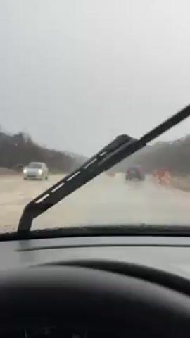 A driver is operating a car on a road that is flooded with muddy water. Rain is falling heavily, and the windshield wipers are in use. Several other cars can be seen in the distance, as well as red and white striped traffic markers. The road is bordered by vegetation on both sides, and the overall visibility is low due to the weather conditions.