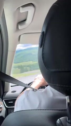 A POV shot from inside a car shows the back of a driver's head and shoulders as they steer. A seatbelt is visible across the driver's chest. Outside the passenger window, a rural landscape with green hills and a cloudy sky can be seen. The car appears to be moving.