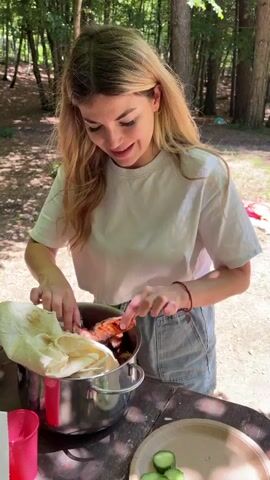 A young woman with long blonde hair, wearing a white t-shirt and denim shorts, stands at a wooden picnic table outdoors. She is positioned in front of a large metal pot containing grilled meat and white flatbread. She uses her right hand to take a piece of meat from the pot. After briefly looking at the meat and making a puckering gesture towards the camera, she places the meat onto a light-colored round plate that already holds several slices of cucumber. Trees and natural light are visible in the background. A red plastic cup is also on the table to her left.